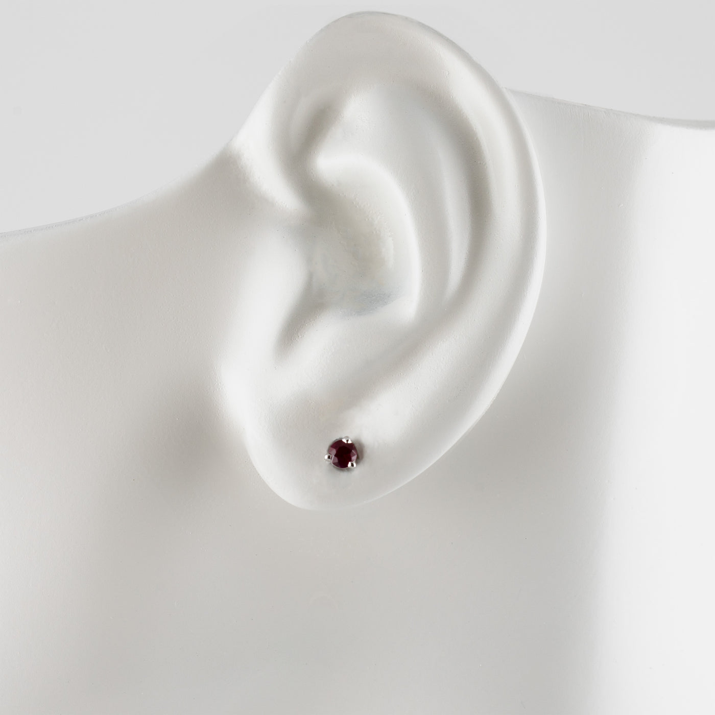 A single small stud earring with a round dark red gemstone, displayed on a white mannequin ear stand against a light gray background.