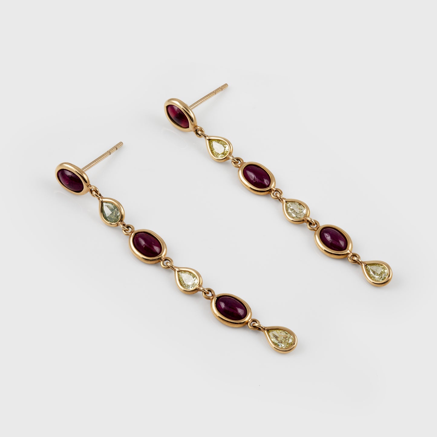 A pair of gold drop earrings featuring oval dark red cabochon stones and teardrop-shaped light yellow or green gemstones, displayed on a clean light gray background.
