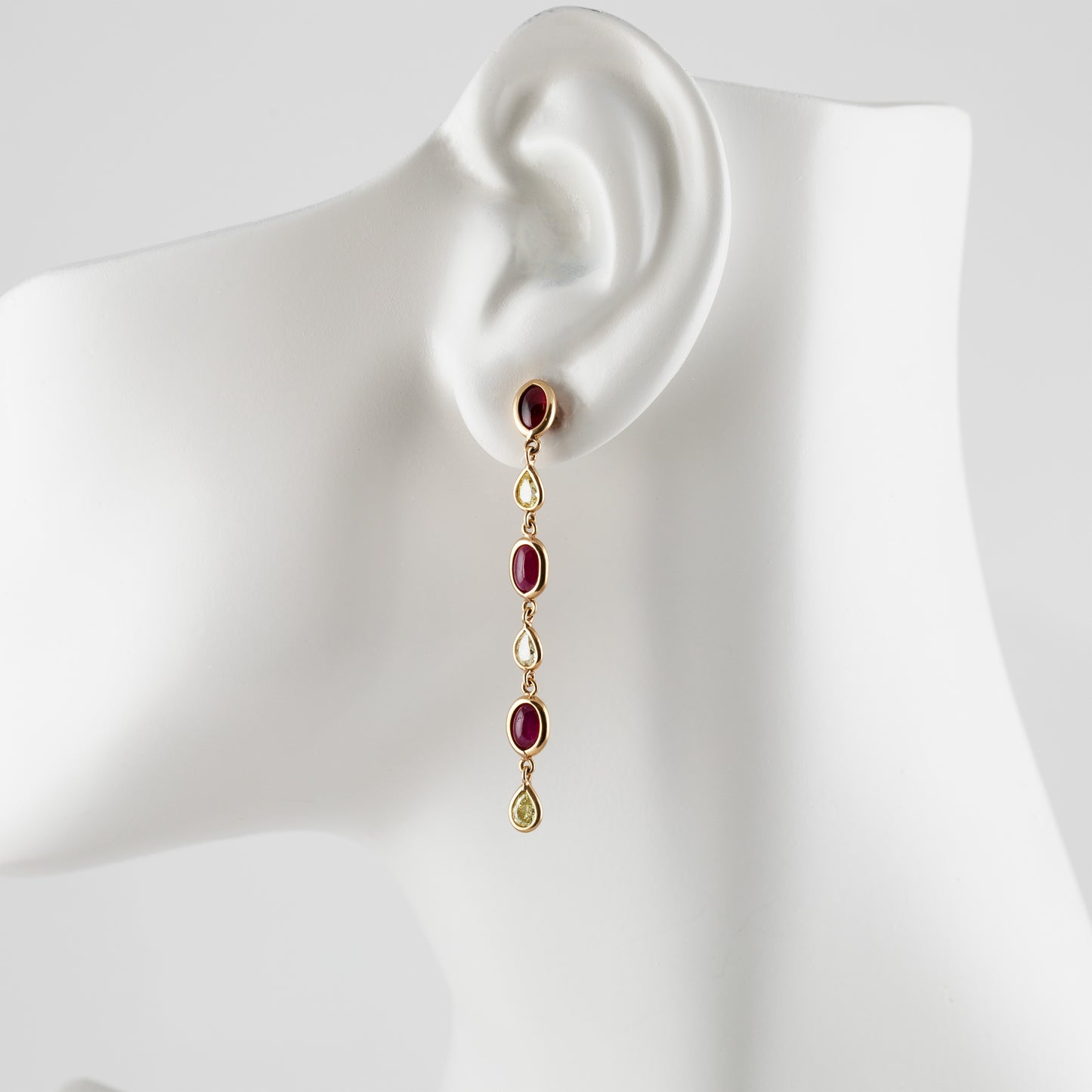 A gold drop earring with oval red gemstones and teardrop-shaped diamonds, displayed on a white mannequin ear against a light gray background.