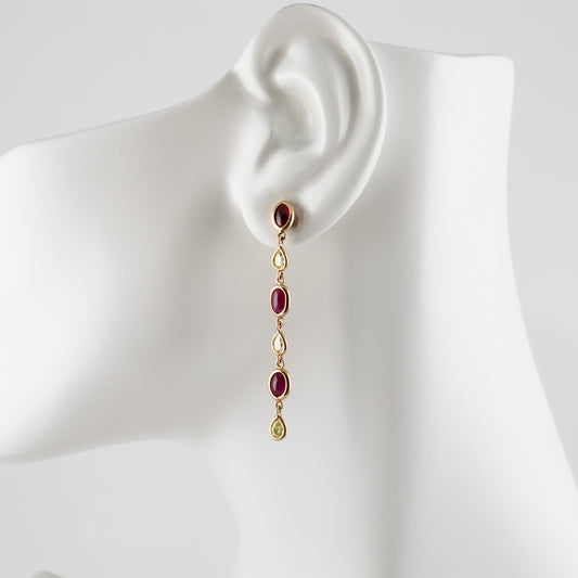 A gold drop earring with oval red gemstones and teardrop-shaped diamonds, displayed on a white mannequin ear against a light gray background.