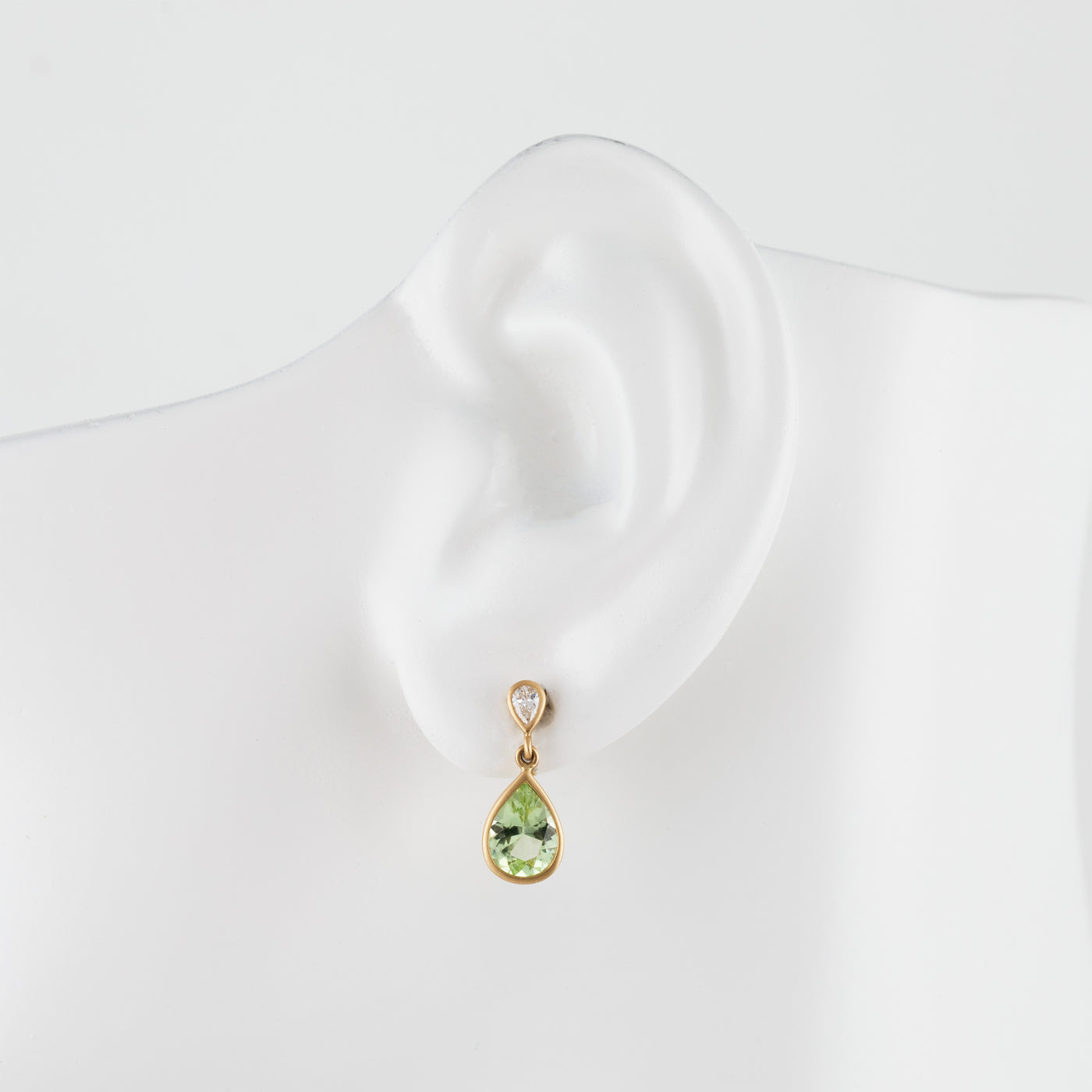A gold drop earring with a light green teardrop gemstone and a small diamond accent stone, displayed on a white mannequin ear.