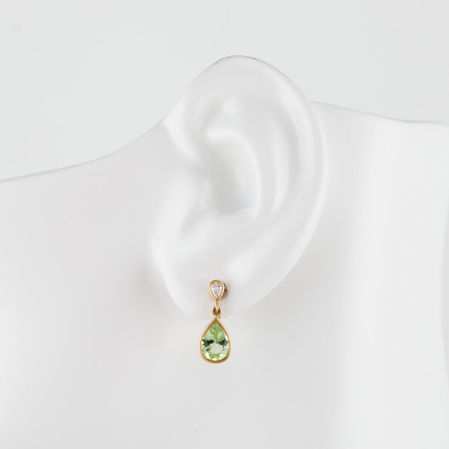 A gold drop earring with a light green teardrop gemstone and a small diamond accent stone, displayed on a white mannequin ear.