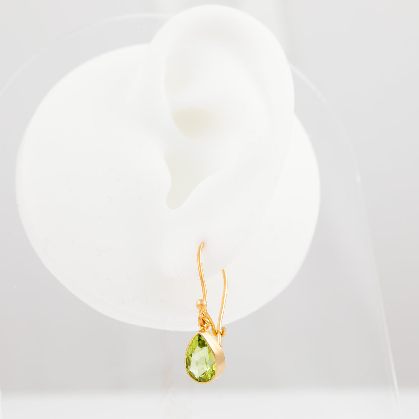 A gold dangle earring with a faceted green teardrop gemstone, displayed on a white ear-shaped jewelry stand against a light gray background.