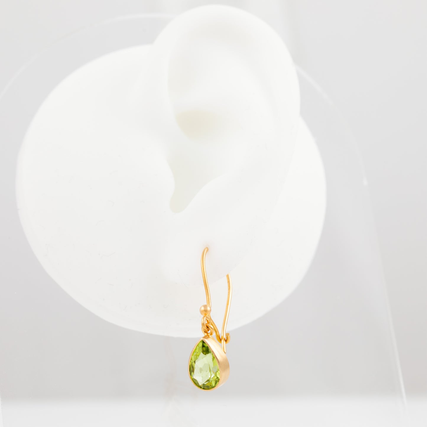 A gold dangle earring with a faceted green teardrop gemstone, displayed on a white ear-shaped jewelry stand against a light gray background.