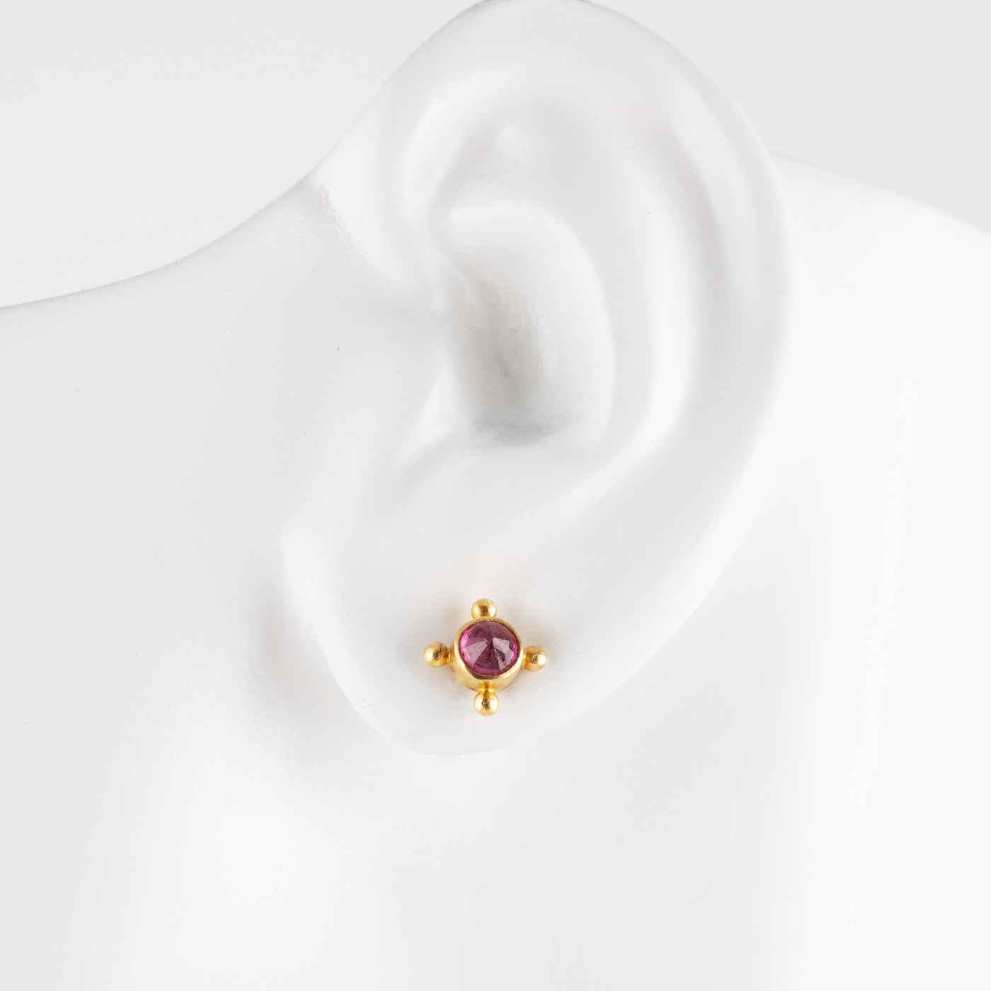 A small gold stud earring with a round gemstone, displayed on a white ear-shaped mannequin stand against a white background.