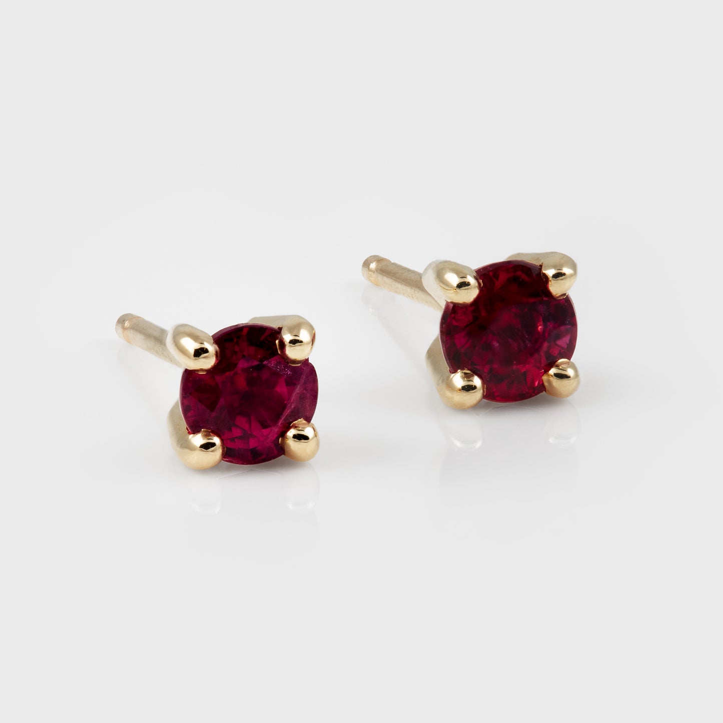 A pair of gold stud earrings with round deep red gemstones, set in a four-prong setting on a light gray reflective background.