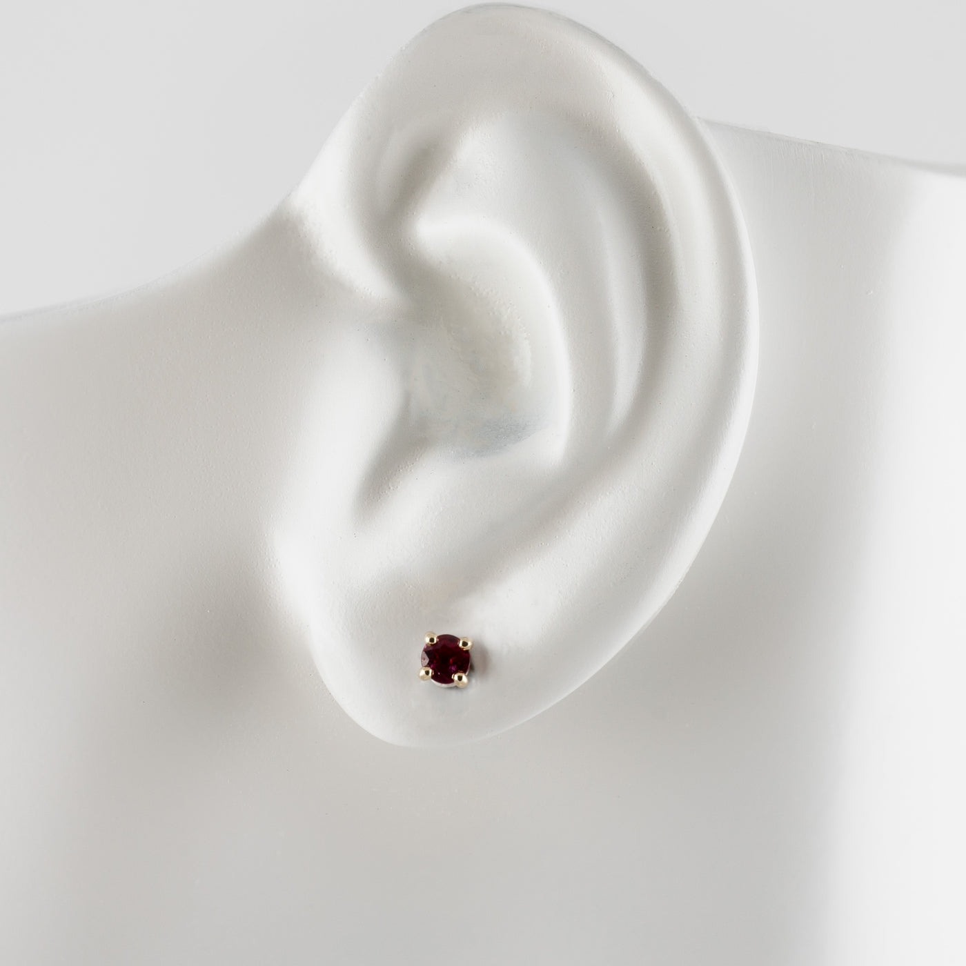 A single stud earring with a small round dark red gemstone in a gold setting, displayed on a white ear-shaped mannequin against a light gray background.