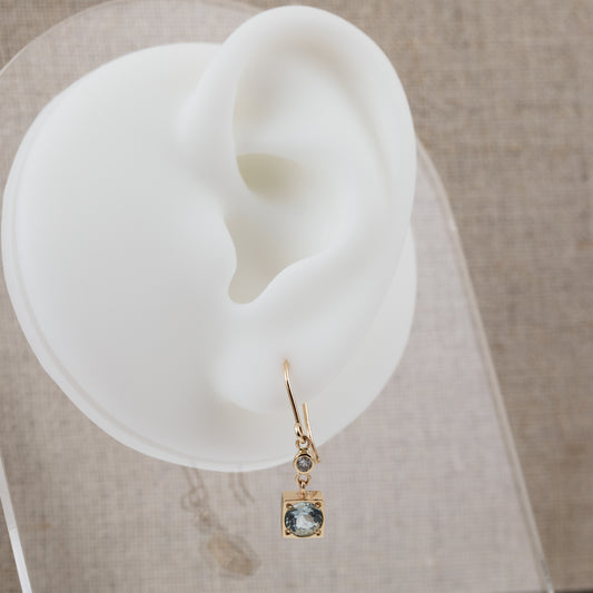 A gold drop earring in a square shape with a light blue aquamarine gemstone and a small round accent stone, displayed on a white silicone ear-shaped stand against a neutral beige textured background