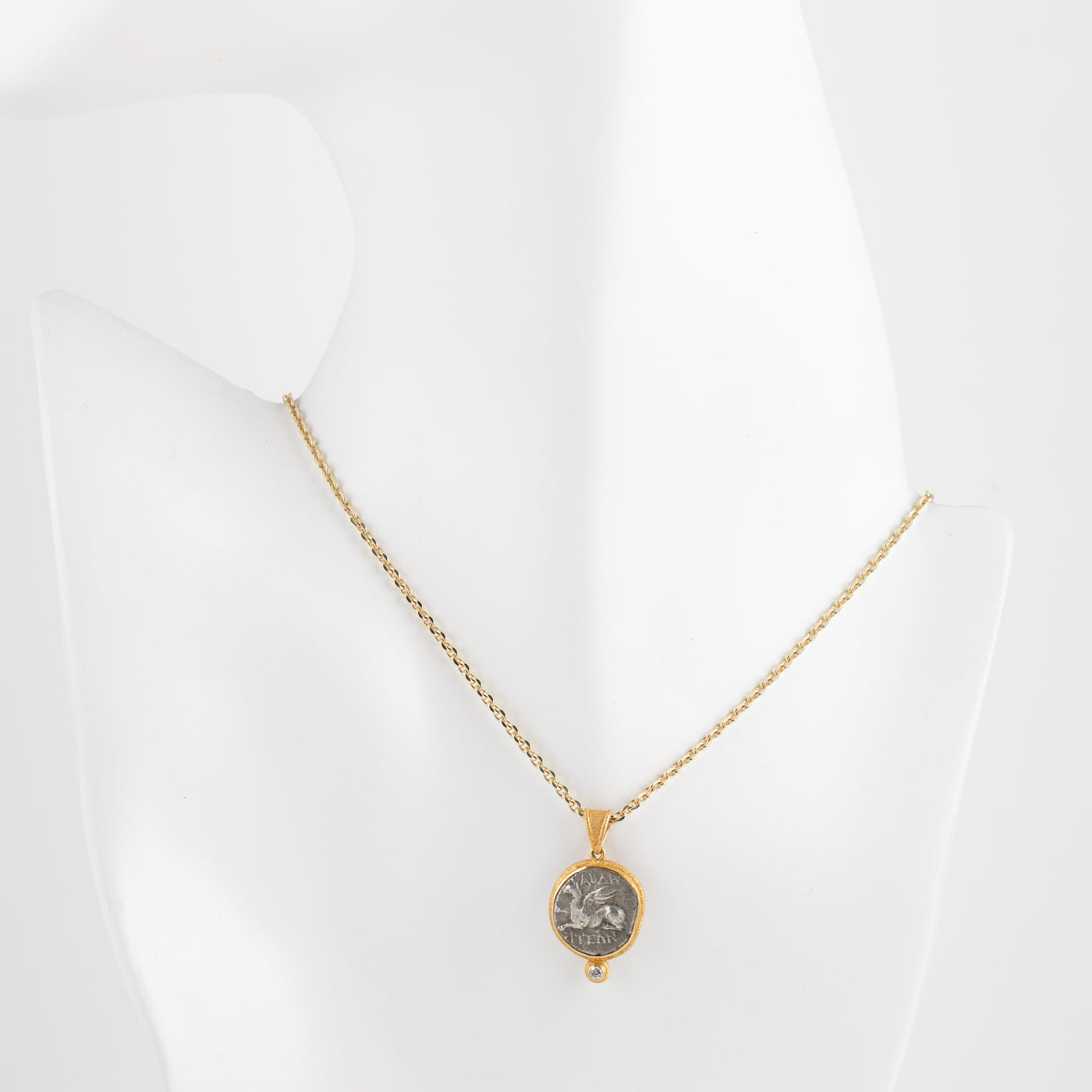 A gold chain necklace with an oval pendant featuring a silver-toned coin, displayed on a white mannequin bust against a white background.