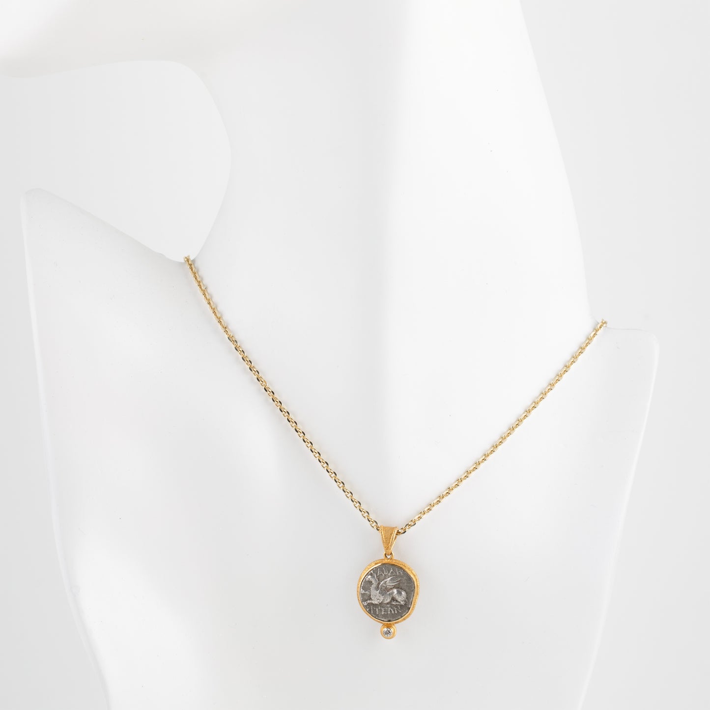 A gold chain necklace with an oval pendant featuring a silver-toned coin, displayed on a white mannequin bust against a white background.