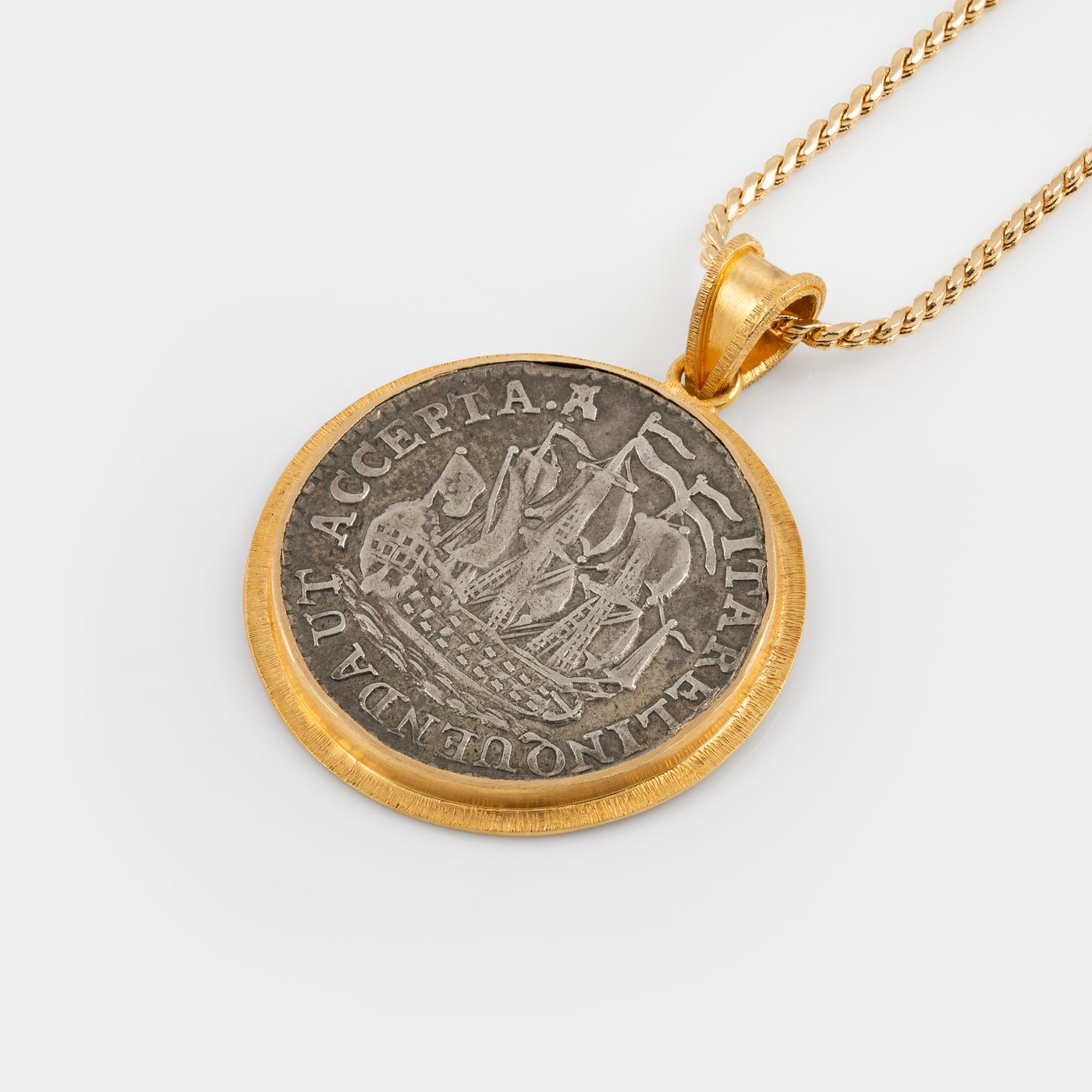 A gold pendant necklace featuring a circular coin pendant with a silver-toned ship design and gold border, suspended from a gold chain against a light gray background.