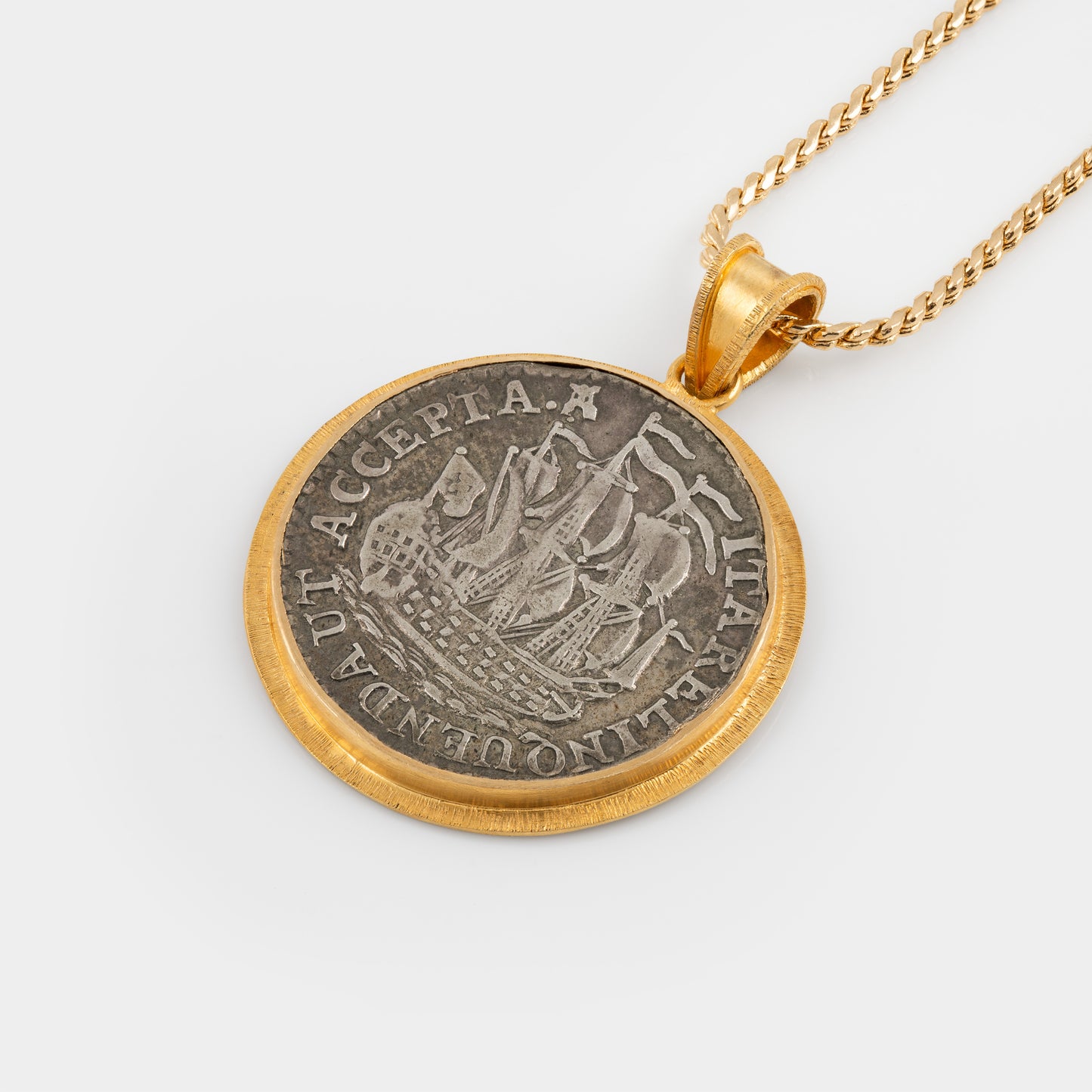 A gold pendant necklace featuring a circular coin pendant with a silver-toned ship design and gold border, suspended from a gold chain against a light gray background.