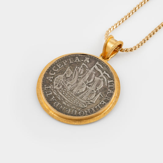 A gold pendant necklace featuring a circular coin pendant with a silver-toned ship design and gold border, suspended from a gold chain against a light gray background.