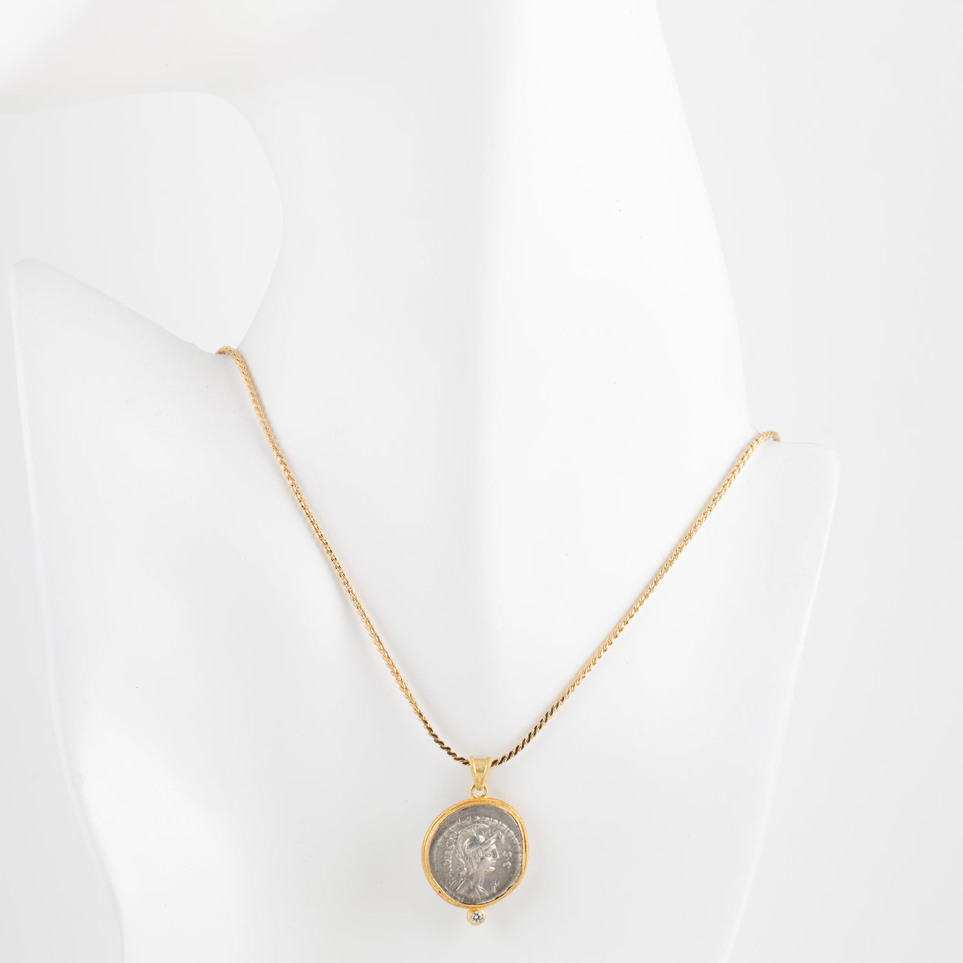 A gold chain necklace with a circular pendant featuring a silver-toned coin, displayed on a white mannequin bust.
