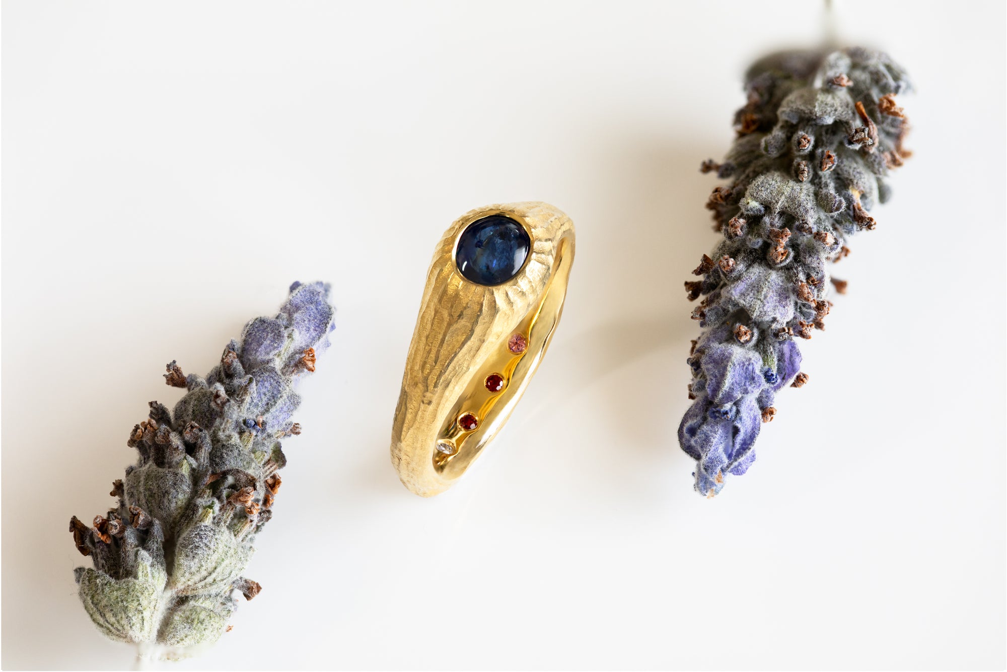 Gold ring with a blue gemstone surrounded by lavender flowers on a white background