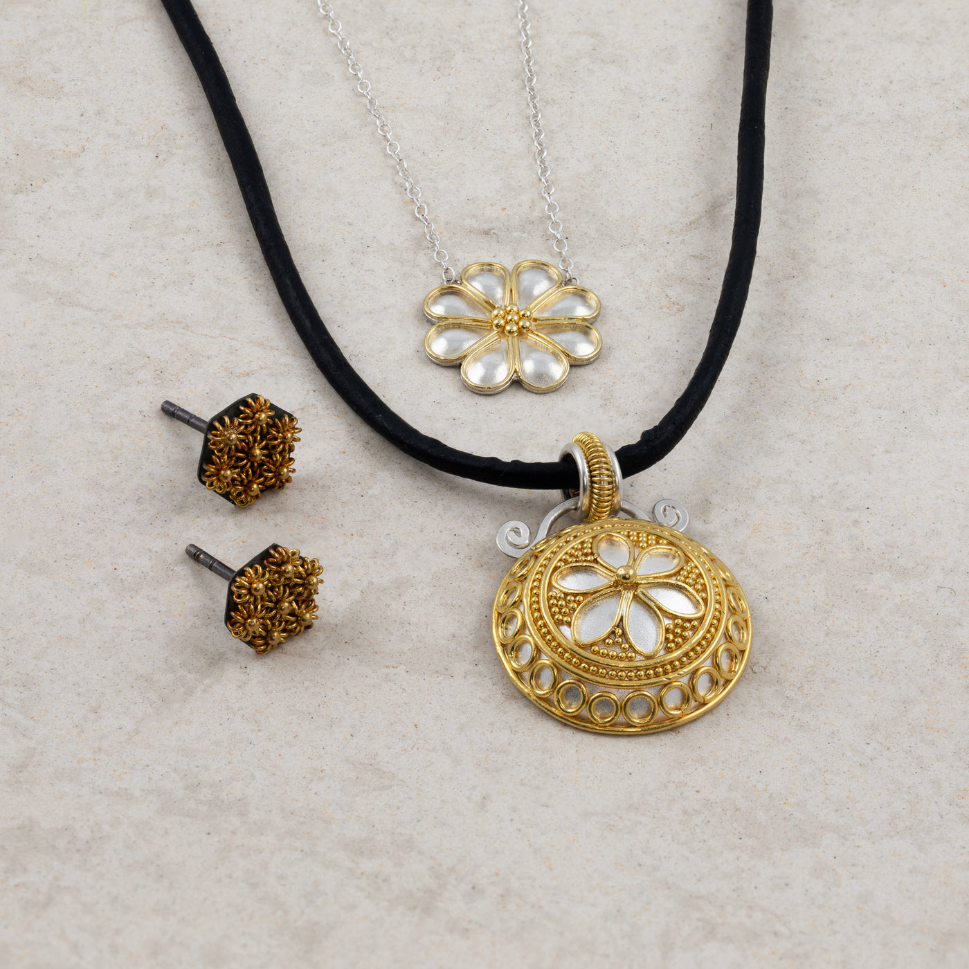Two gold and silver floral pendant necklaces and a pair of stud earrings on a stone background