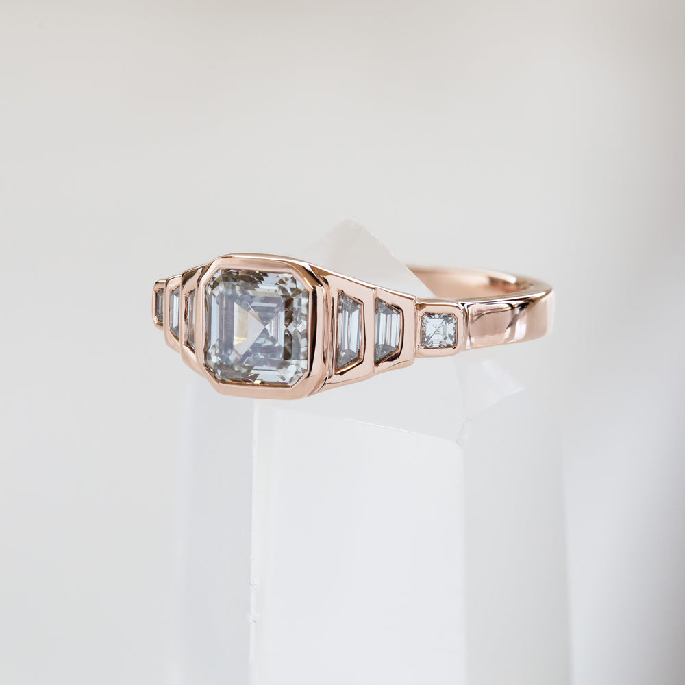 Rose gold ring with tiered diamonds on a white background, balancing on a clear crystal. 