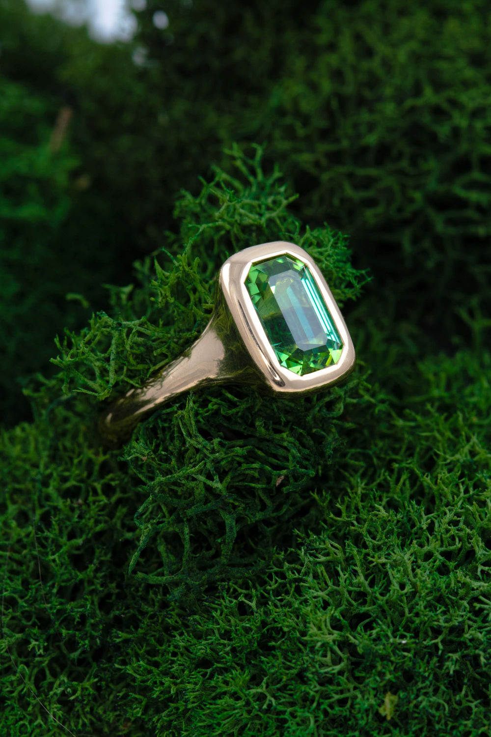 Yellow gold ring featuring emerald cut green gemstone featured against green moss background
