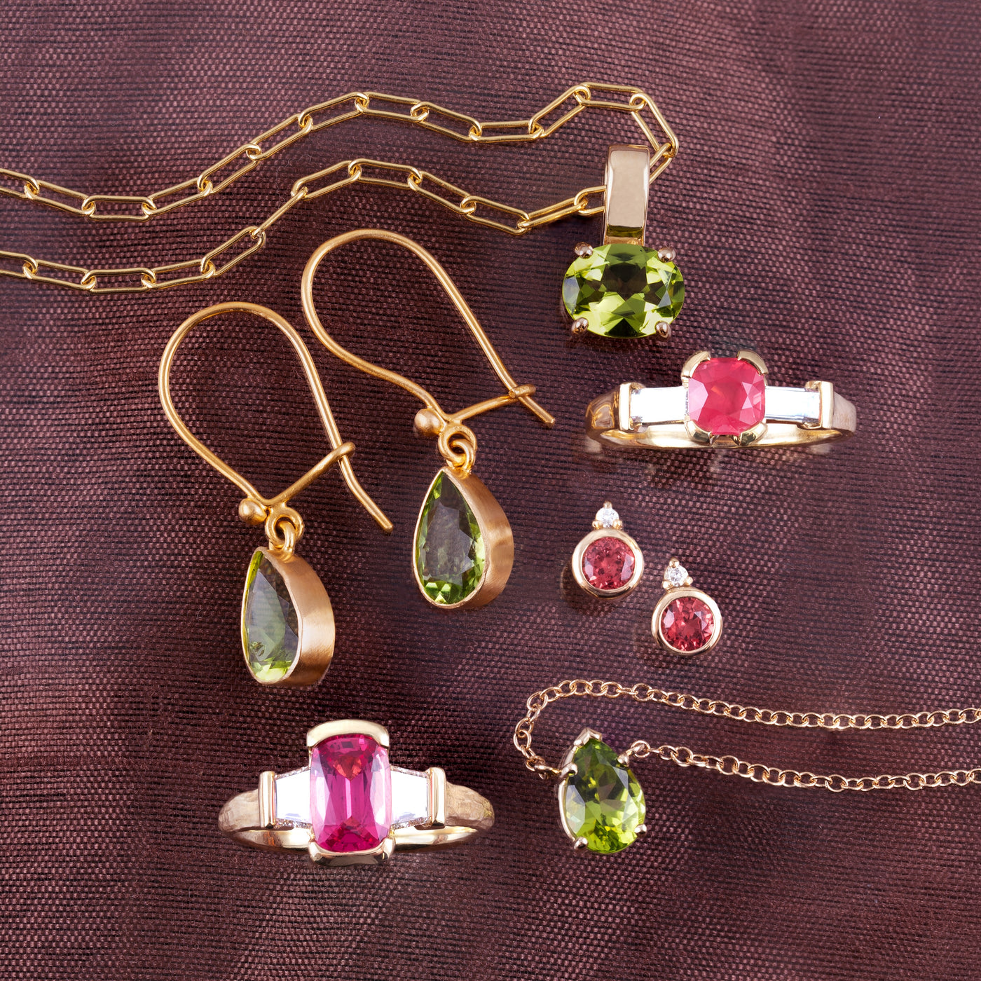 A gold jewelry set featuring a pendant, earrings, rings, and necklaces with green and pink gemstones, arranged on a textured dark brown fabric background.
