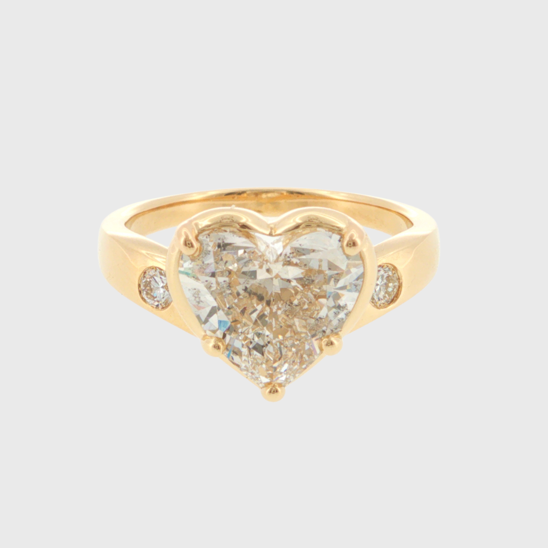 A yellow gold heart-shaped diamond ring with two small round diamonds on the band, set against a light gray background.