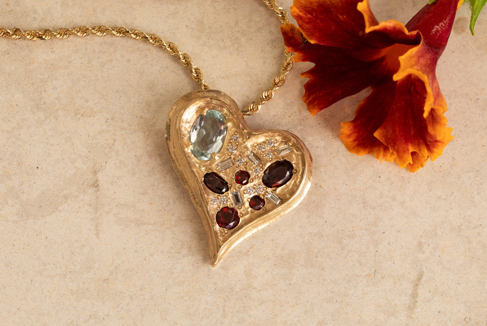 Yellow gold pendant featuring assorted blue, red, and white gems, in the shape of a heart, suspended from chain against textured background with flower accent