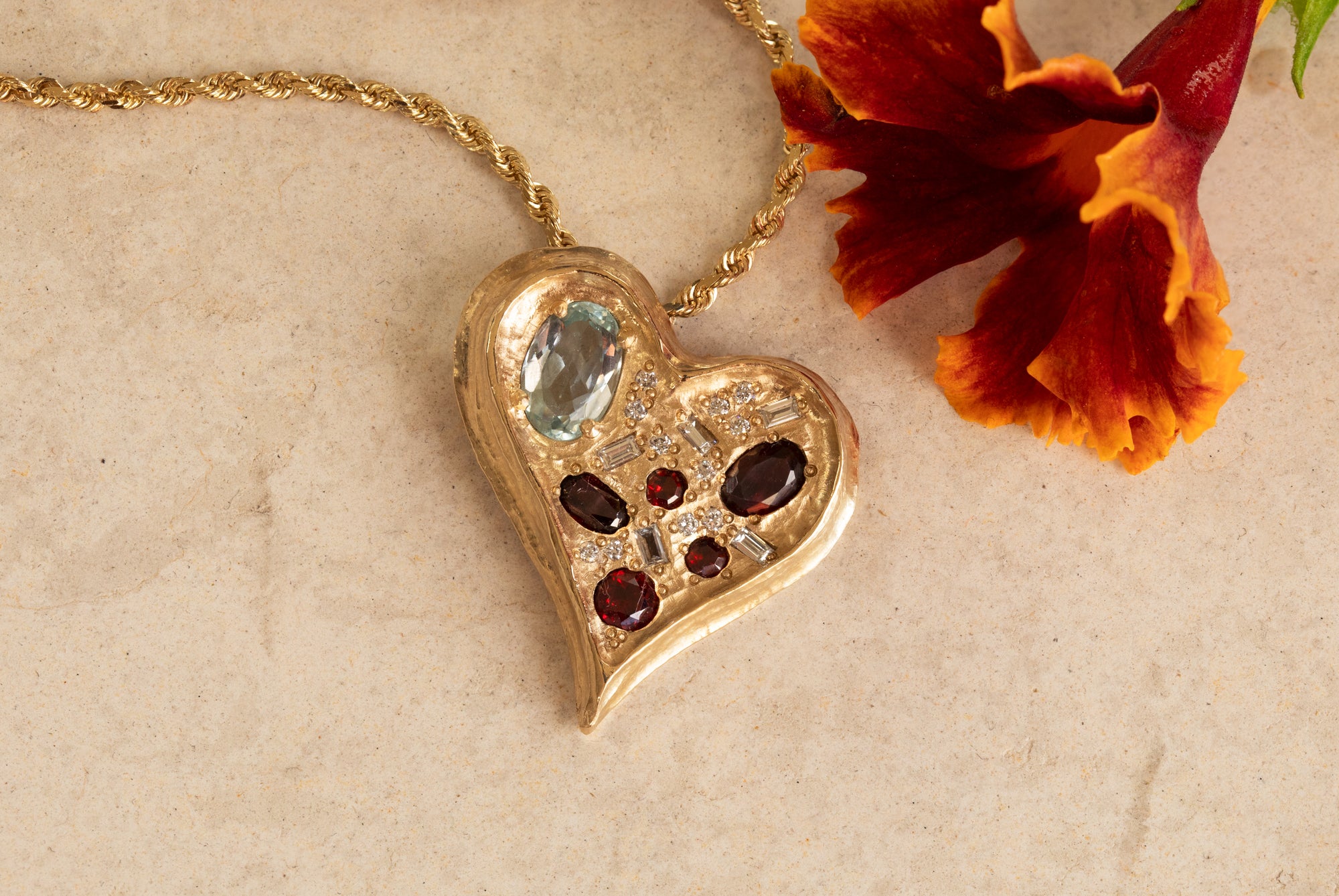 Yellow gold pendant featuring assorted blue, red, and white gems, in the shape of a heart, suspended from chain against textured background with flower accent