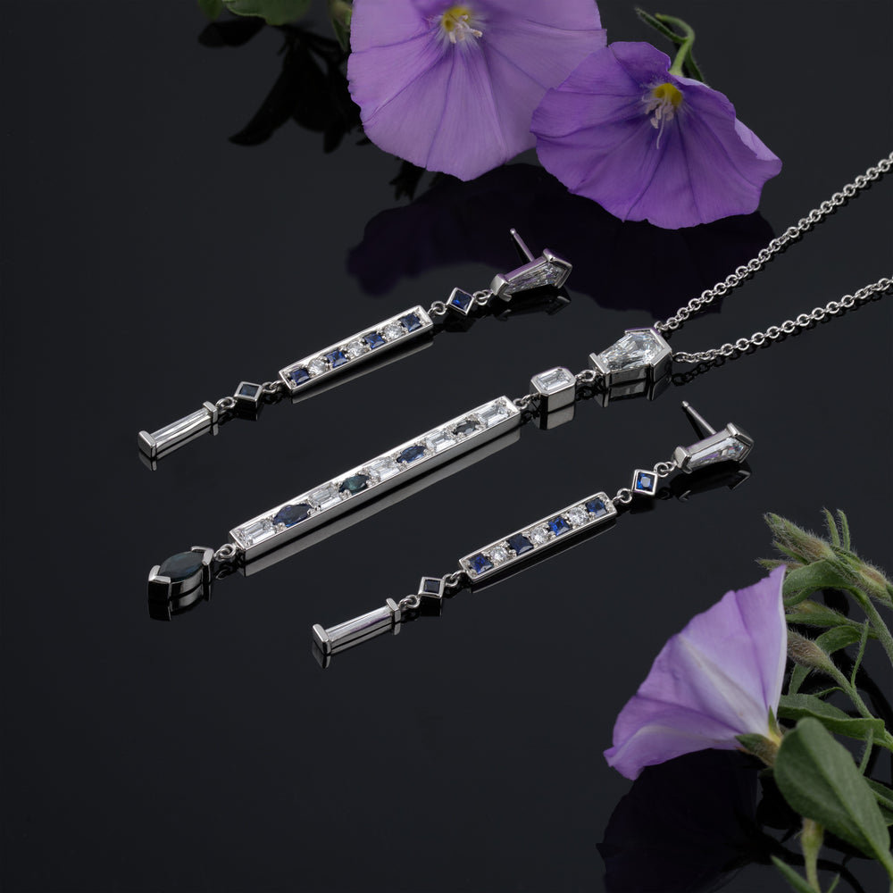 Matching pair of earrings and necklace with hexagonal diamonds at the top, long and skinny with blue gemstones and diamonds along the length, on a reflective black background with purple flowers. 