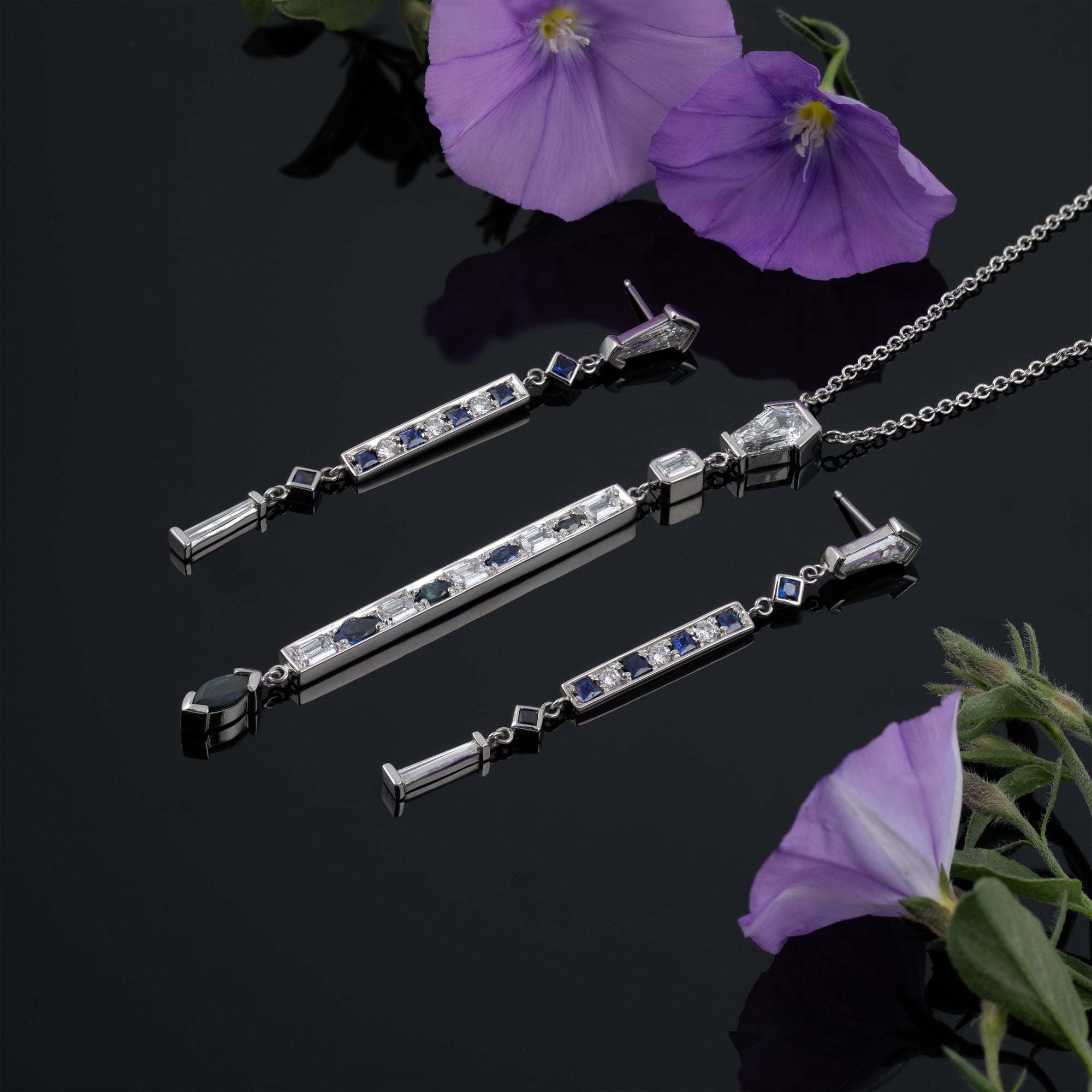 Matching pair of earrings and necklace with hexagonal diamonds at the top, long and skinny with blue gemstones and diamonds along the length, on a reflective black background with purple flowers. 