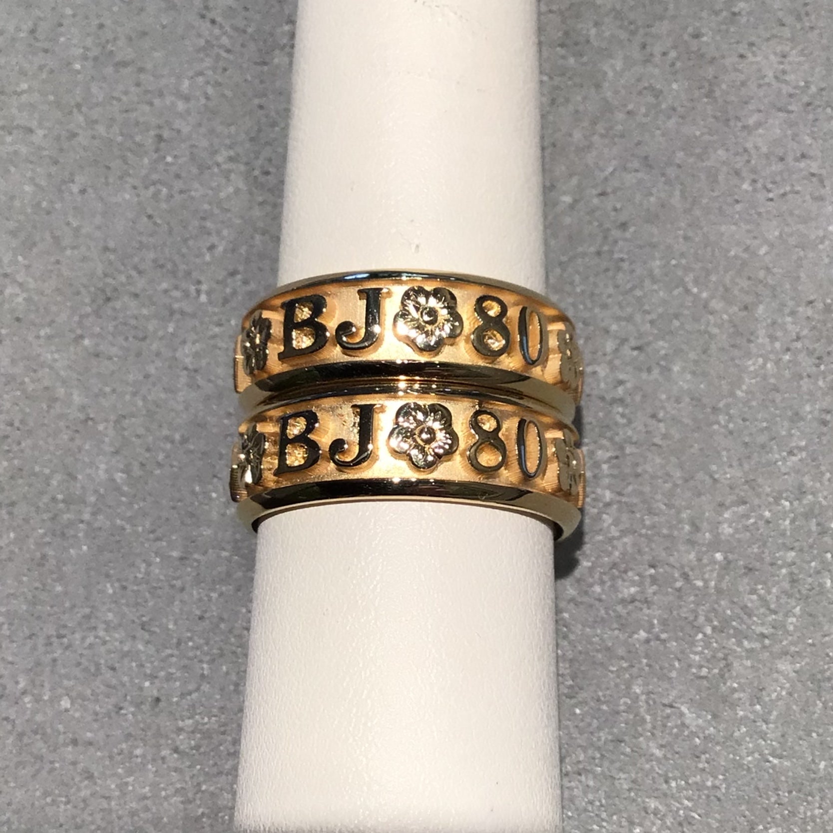 Two bands on a white ring mandrel with a grey background. Rings have the letters BJ, a flower, and 80 on them. 