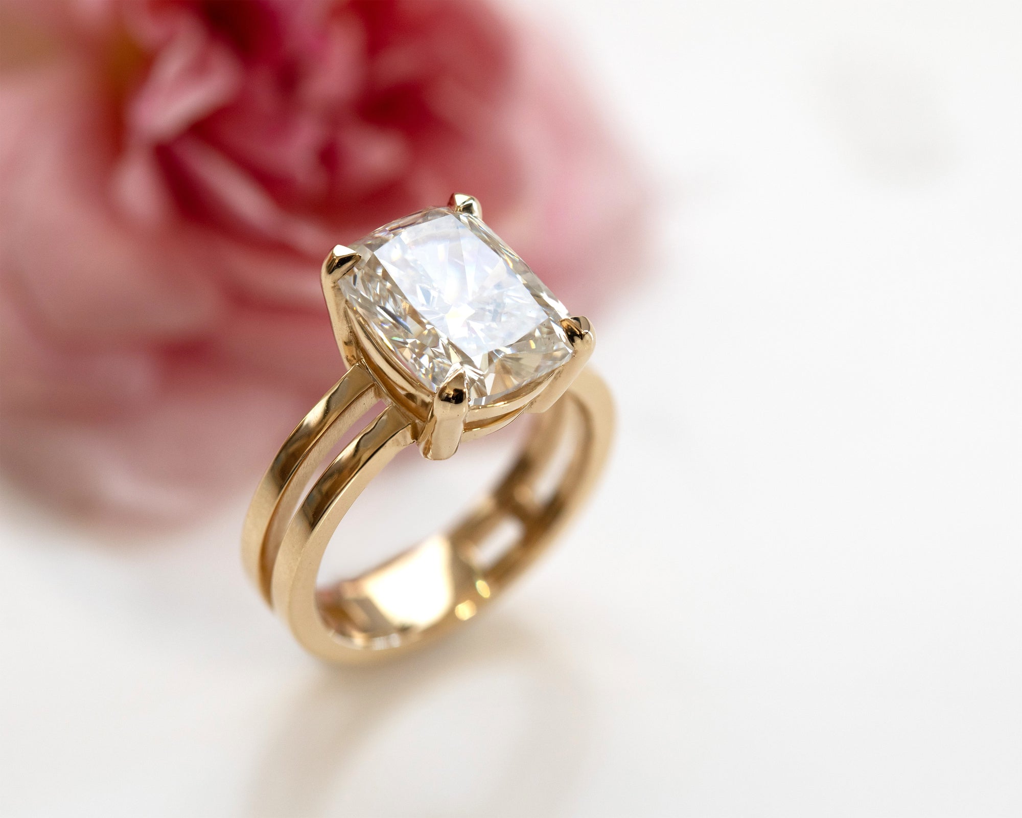 Gold ring with center diamond white background with a pink flower in the upper left corner of the image. 