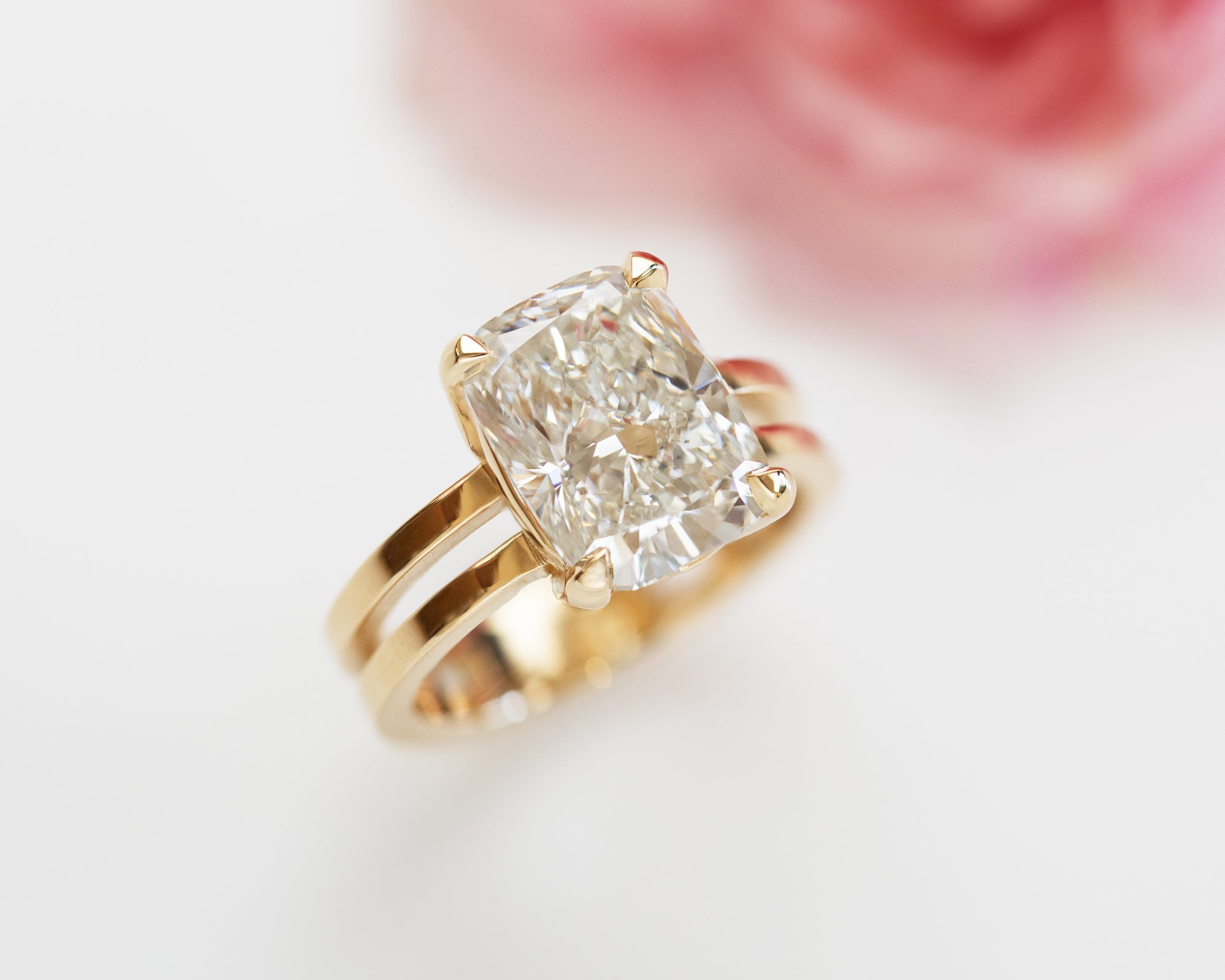 Gold ring with center diamond white background with a pink flower in the upper right corner of the image. 
