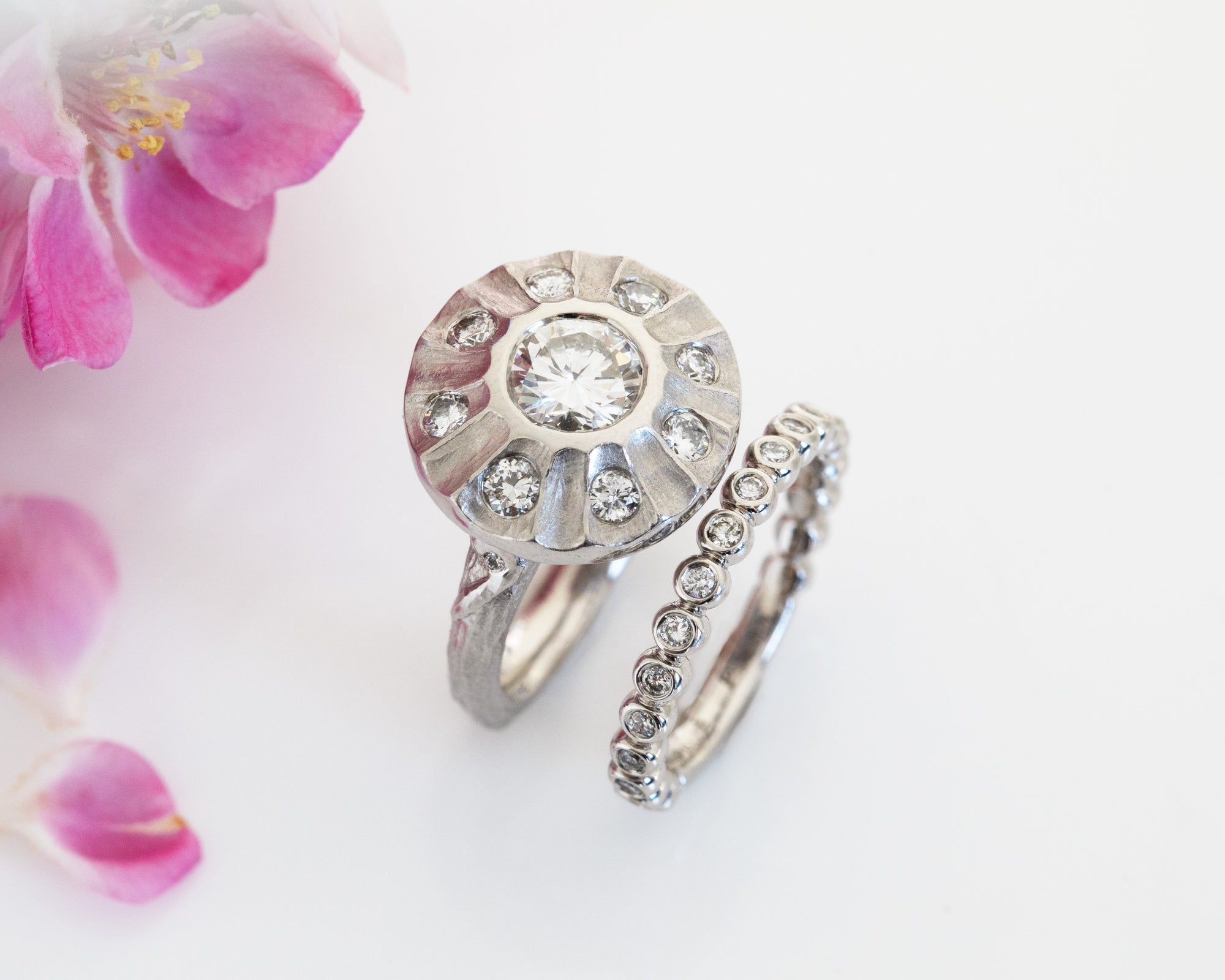Platinum ring with center diamond and accent diamonds in a halo with a diamond eternity band on a white background with pink flowers in the left corner of the image. 