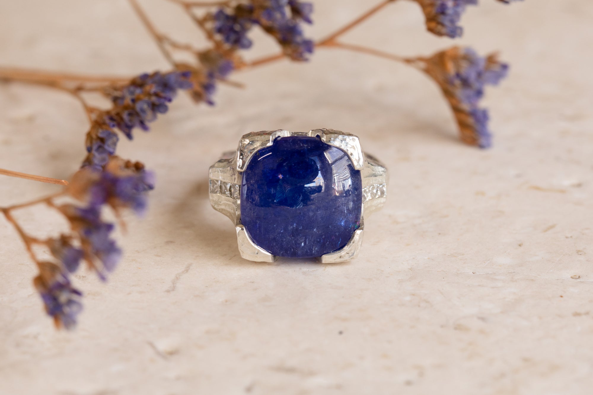 A silver ring featuring a large, cushion-cut deep blue tanzanite, styled with dried purple flowers on a textured beige stone surface.