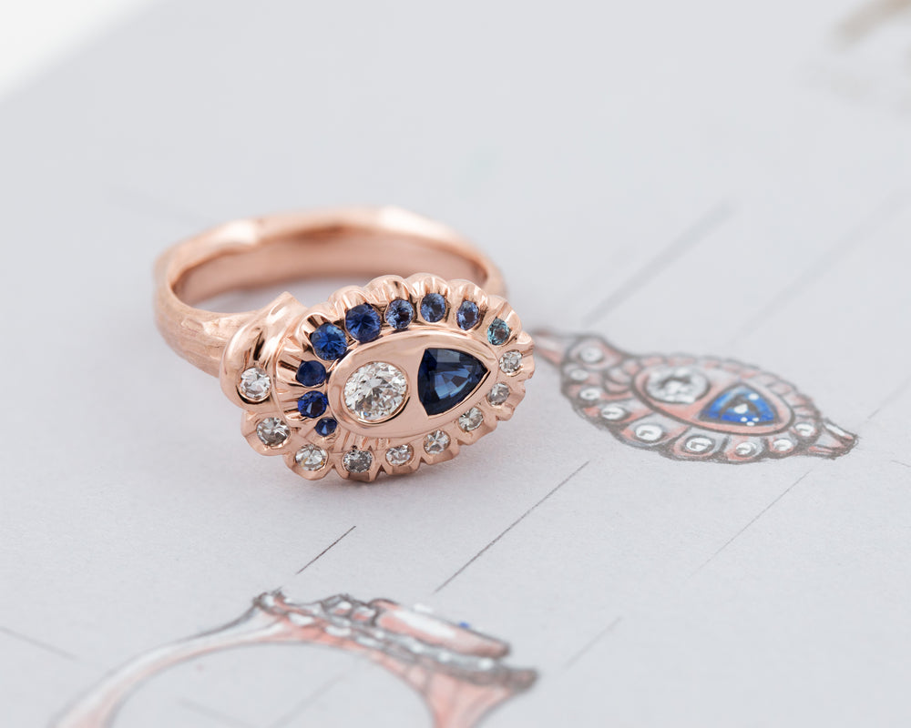 Rose gold ring with a spiral pattern of diamonds and blue gemstones on a grey background with sketches