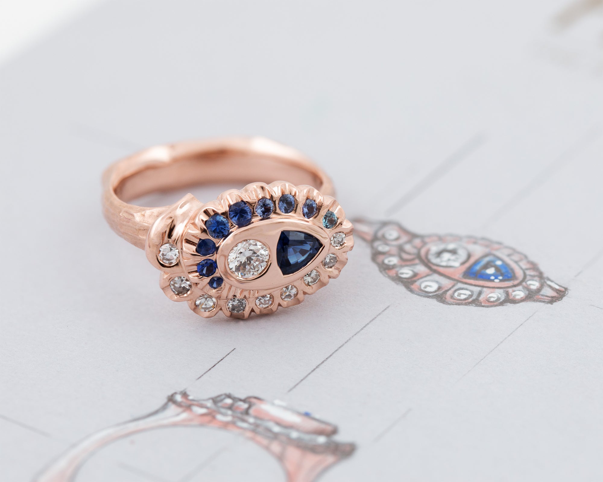 Rose gold ring with a spiral pattern of diamonds and blue gemstones on a grey background with sketches