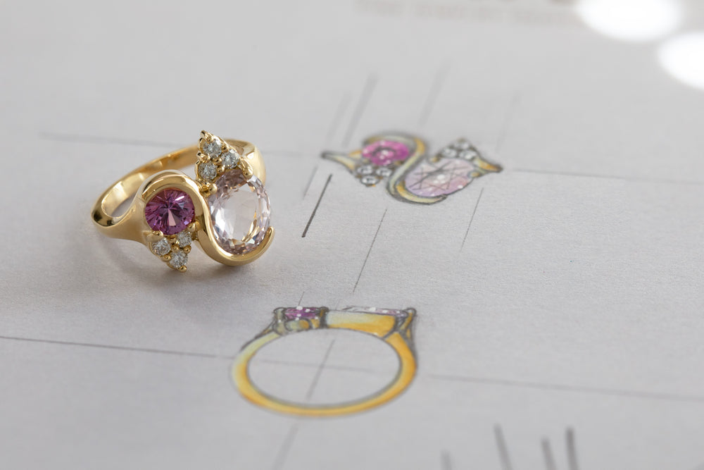 Gold ring with a various white and purple gemstones, photographed on a light gray surface with faint sketch in the background.