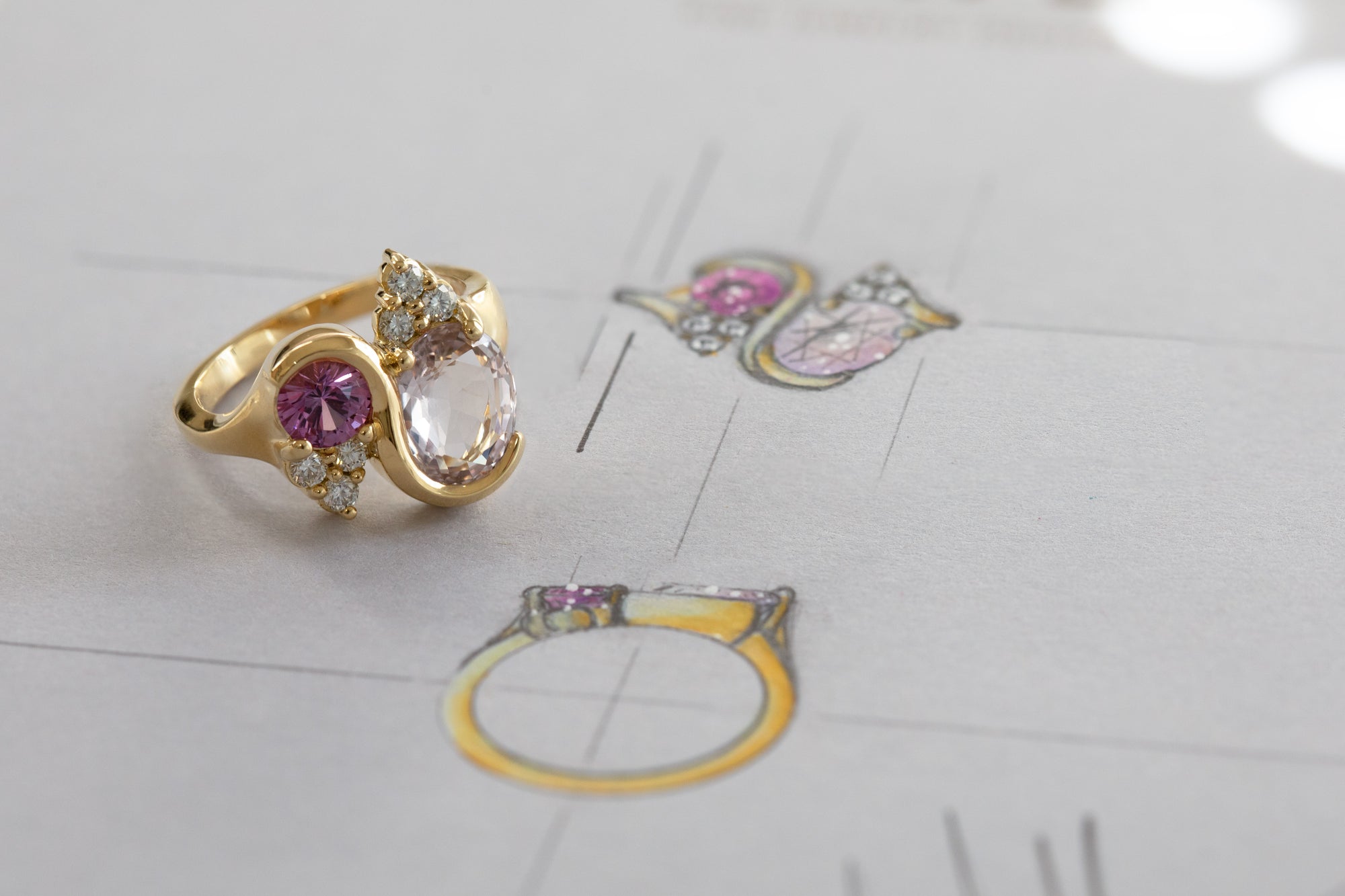 Gold ring with a various white and purple gemstones, photographed on a light gray surface with faint sketch in the background.