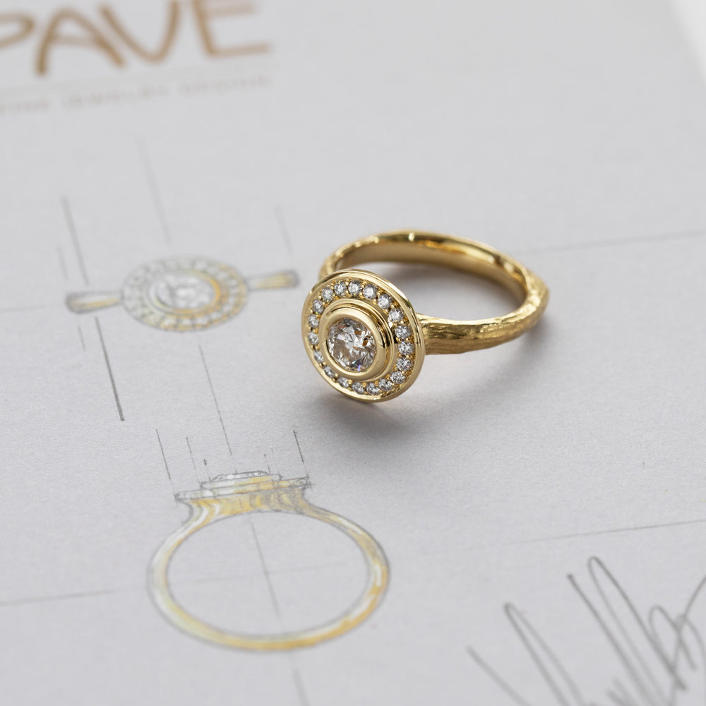 Yellow gold ring featuring bezel set round Diamond with halo of accent diamonds against background of design sketch