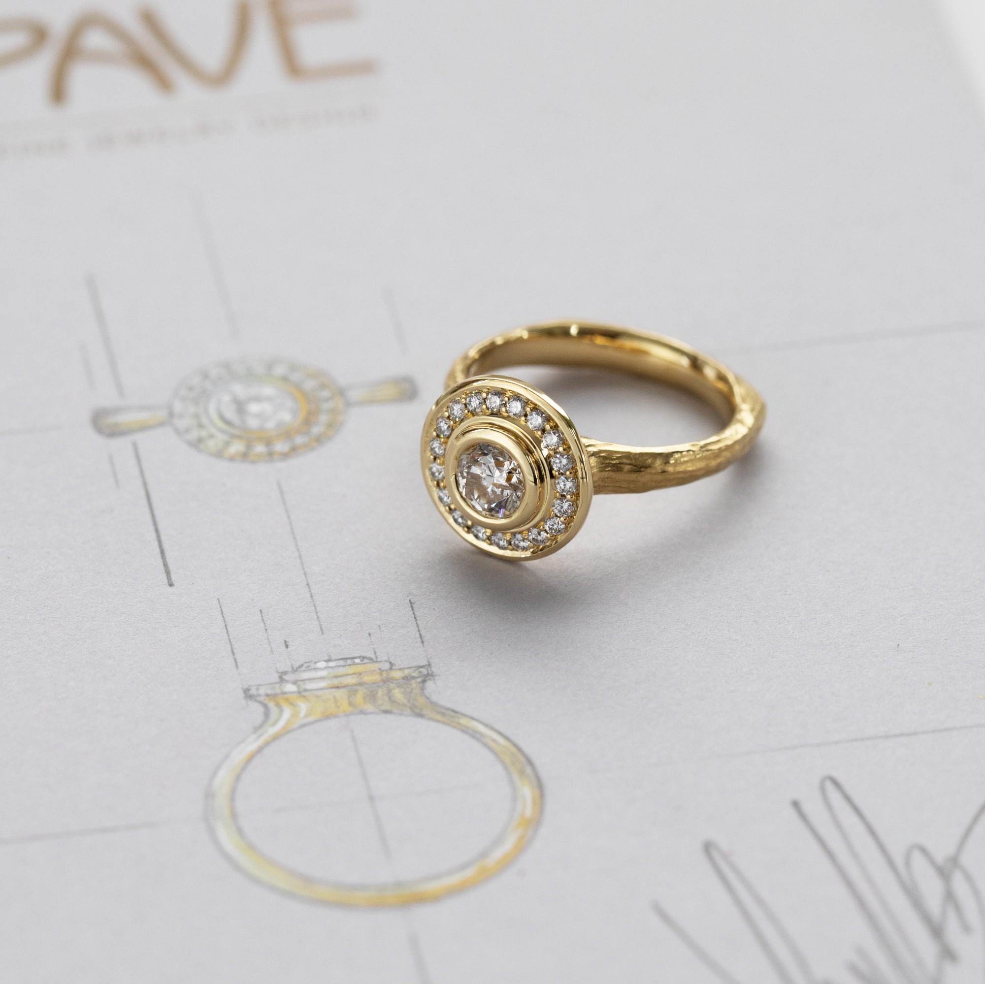 Yellow gold ring featuring bezel set round Diamond with halo of accent diamonds against background of design sketch