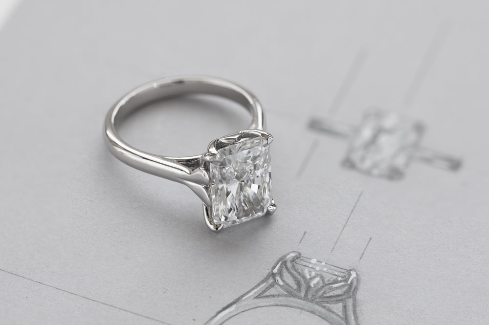 A white metal ring with a rectangular diamond, photographed on a light gray surface with faint sketch in the background.