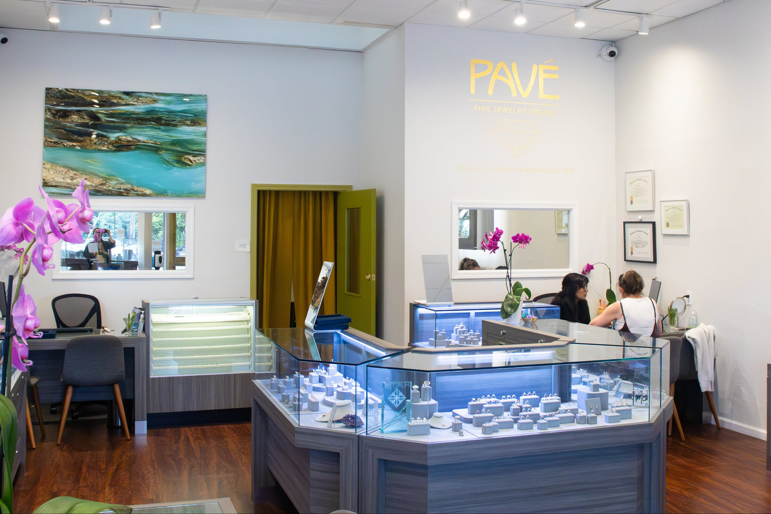 A jewelry showroom interior with multiple glass display cases, wood flooring, track lighting, and a yellow wall logo reading PAVÉ. Several people are seated or standing near the back of the room, and decorative pink flowers and framed wall art are visible.