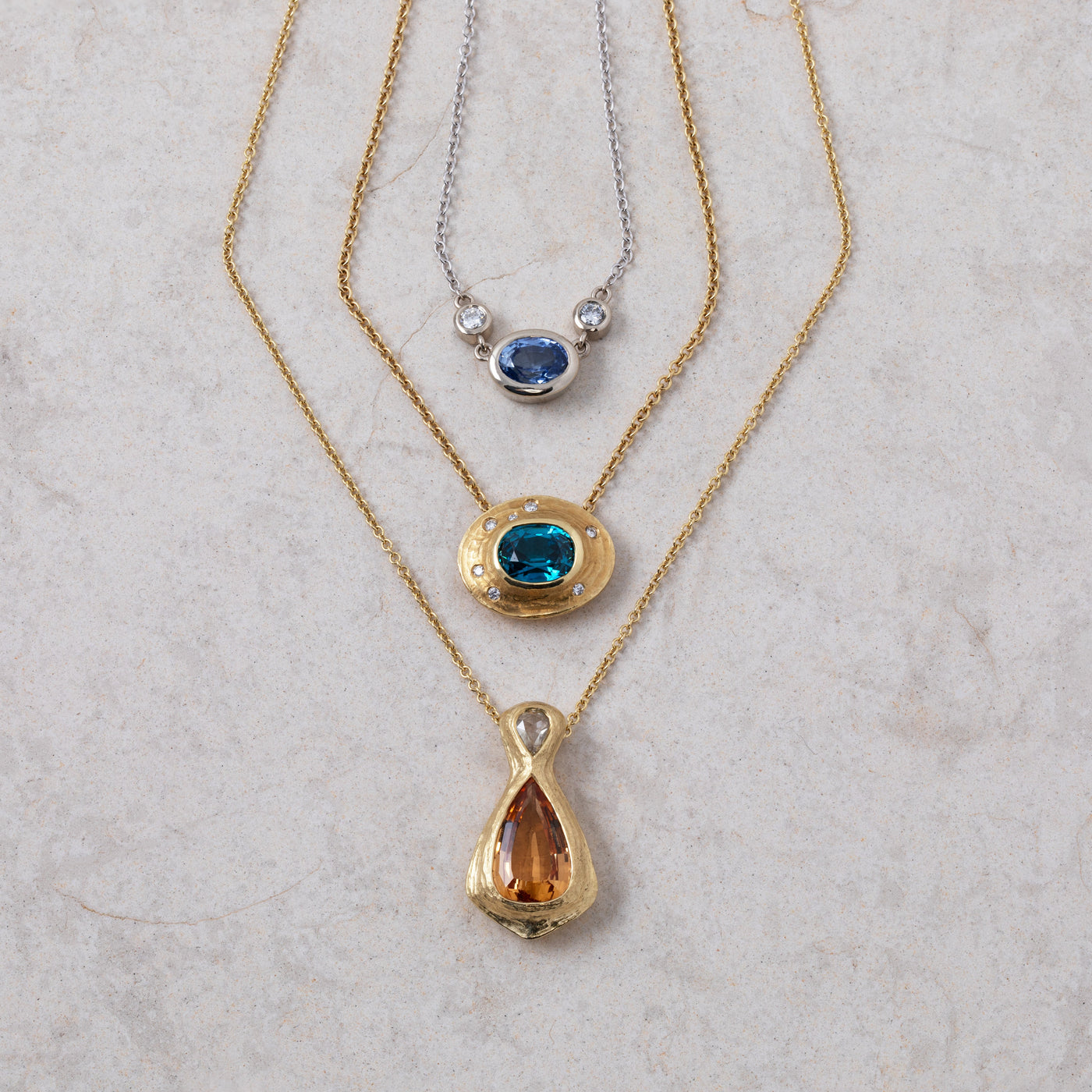Three necklaces on a pale tan slate background, one gold with an orange topaz gemstone, one with a blue zircon gemstone, and one with a blue sapphire gemstone, all with small accent diamonds 