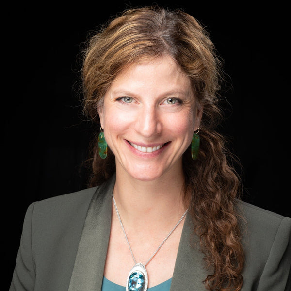 Woman wearing a green blazer and blue top with a necklace against a black background