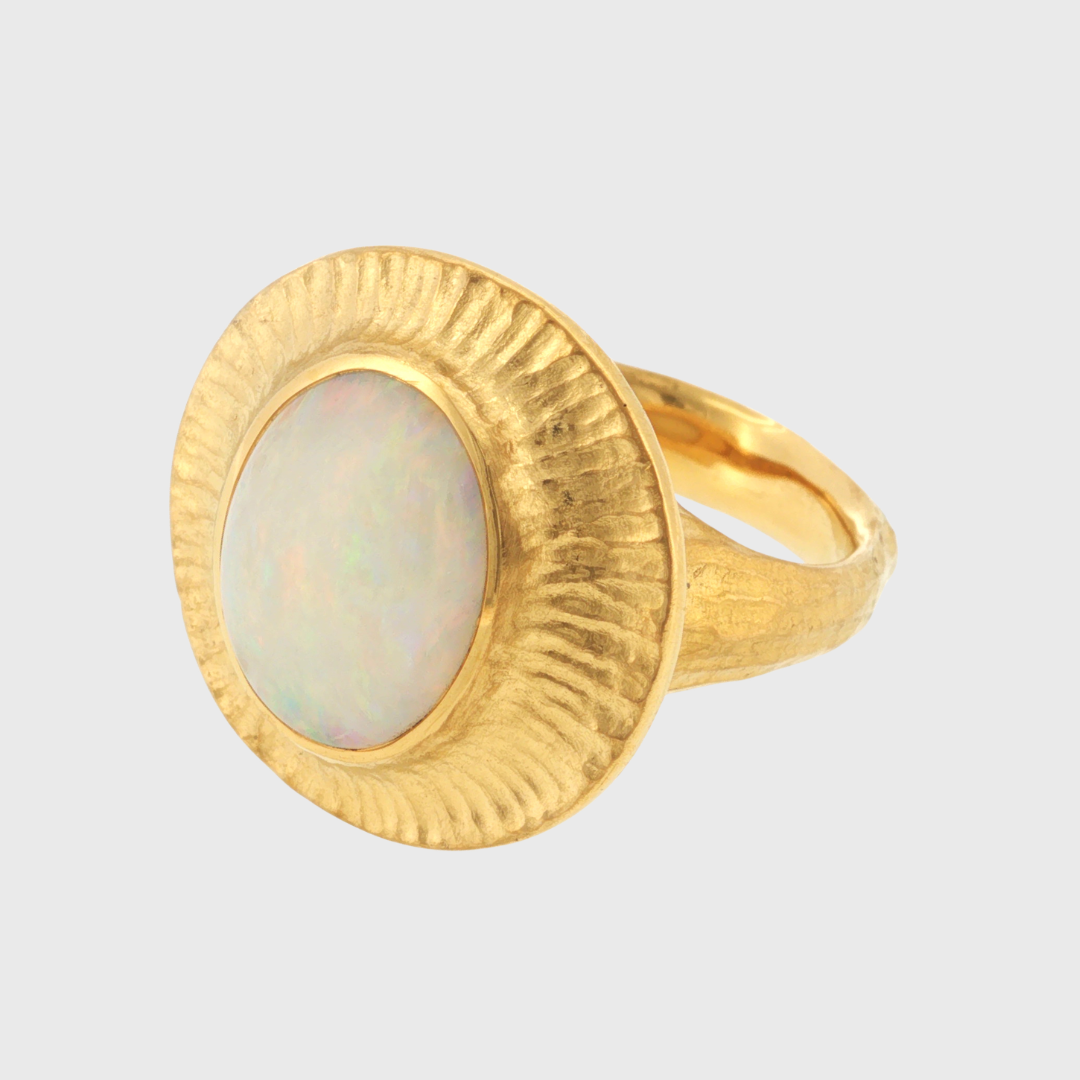 A textured gold ring featuring a large, oval-shaped white opal stone in a wide, sunburst-style bezel setting, shown against a light gray background.