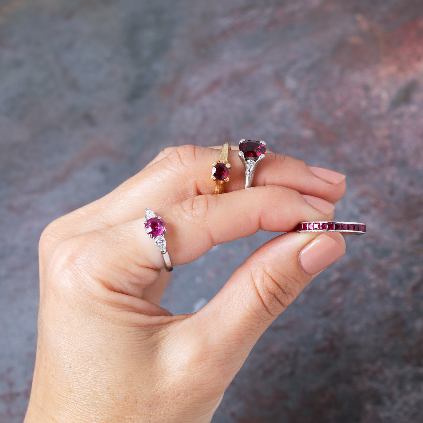 A hand holds several rings against a blurred gray-brown background. The rings include platinum and gold bands with deep red gemstones
