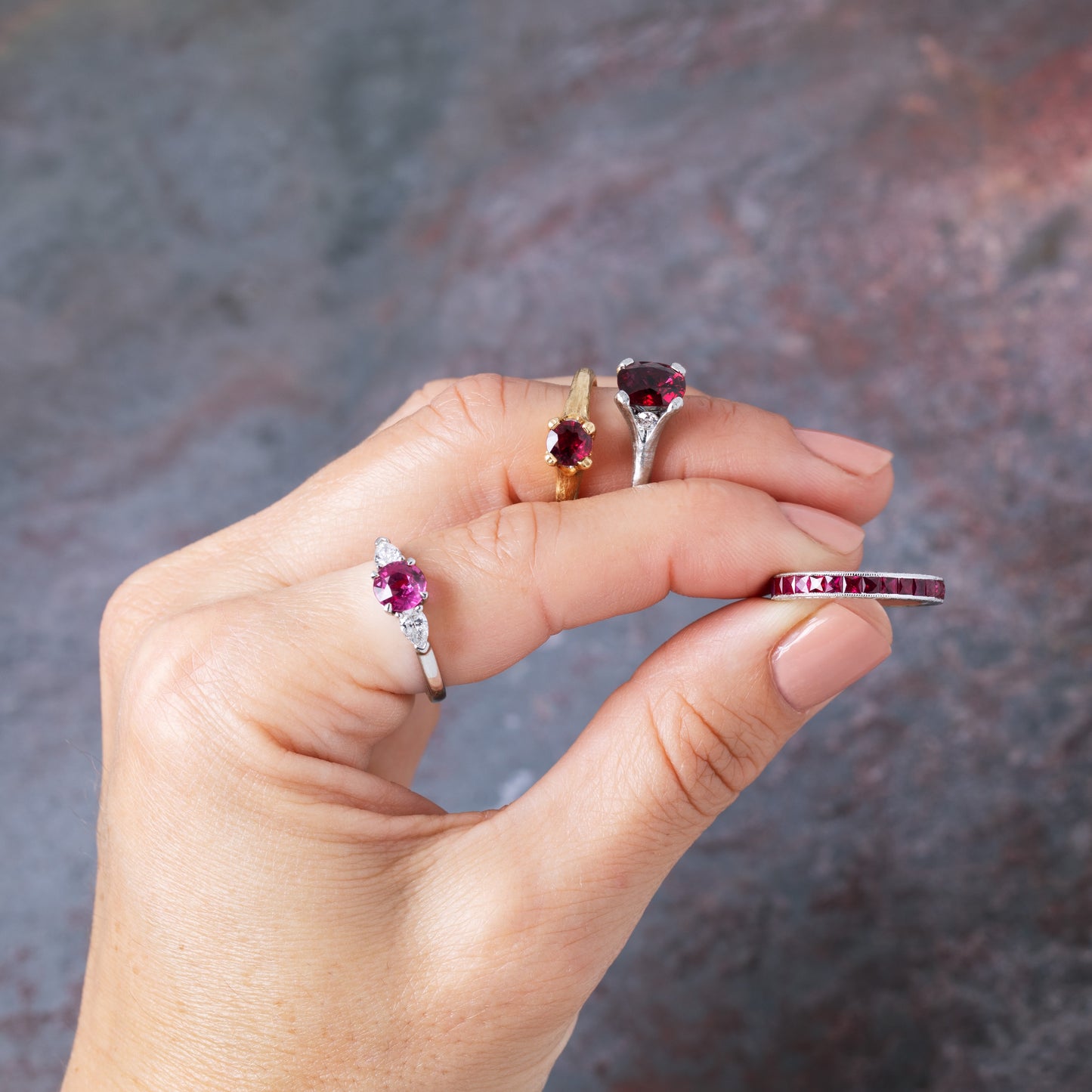 A hand holds several rings against a blurred gray-brown background. The rings include platinum and gold bands with deep red gemstones